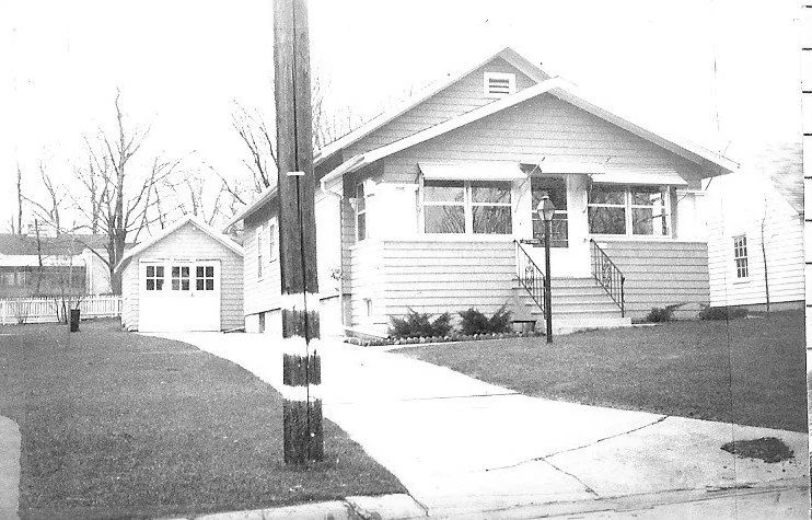 Black-and-white photo of a single-story bungalow with a front porch and steps, detached garage at the end of a curved driveway, and a utility pole in the foreground.