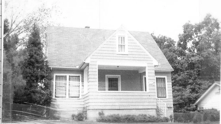 Black-and-white photo of a small wood-sided single-family house with a gabled roof, covered front porch, and trees on either side.