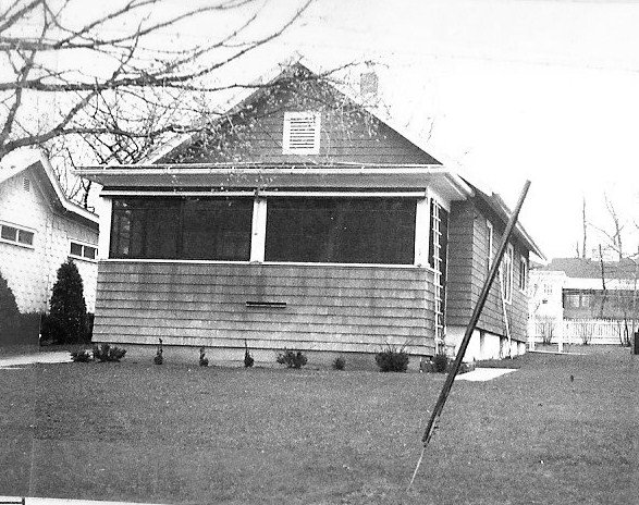 Black-and-white photo of a single-story bungalow with an enclosed front porch, small shrubs along the foundation, and a leaning utility pole in the front yard.