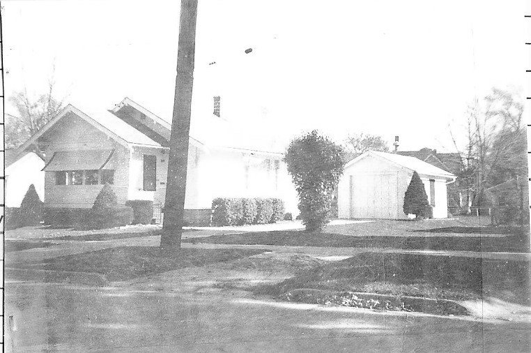 Black-and-white photo of a bungalow-style house with a detached garage, trimmed shrubs and trees, and a utility pole in the front yard.