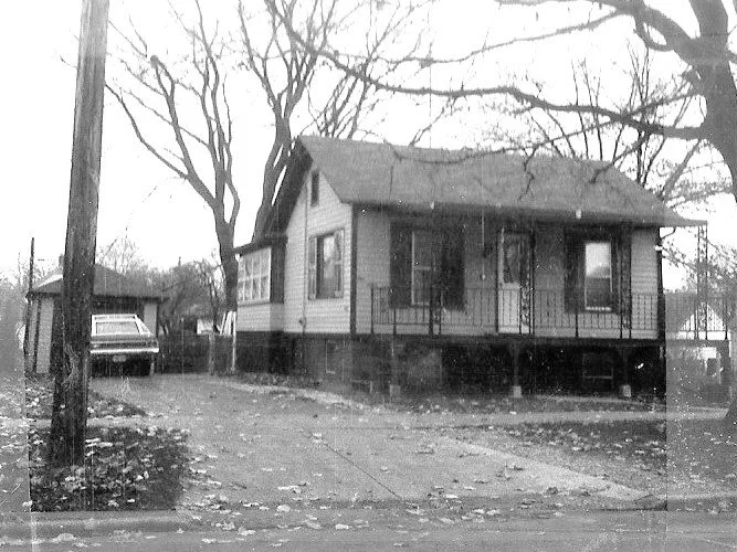 Black-and-white photo of a small raised bungalow with a front porch, bare trees, driveway and an older car parked to the left.