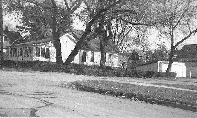 Black-and-white photo of a single-story house with a screened porch, large trees in front, a detached garage to the right, and a cracked street in the foreground.