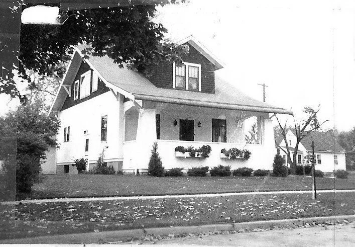 Black-and-white photo of a two-story Craftsman-style house with a covered front porch, window flower boxes, and a lawn with sidewalk.