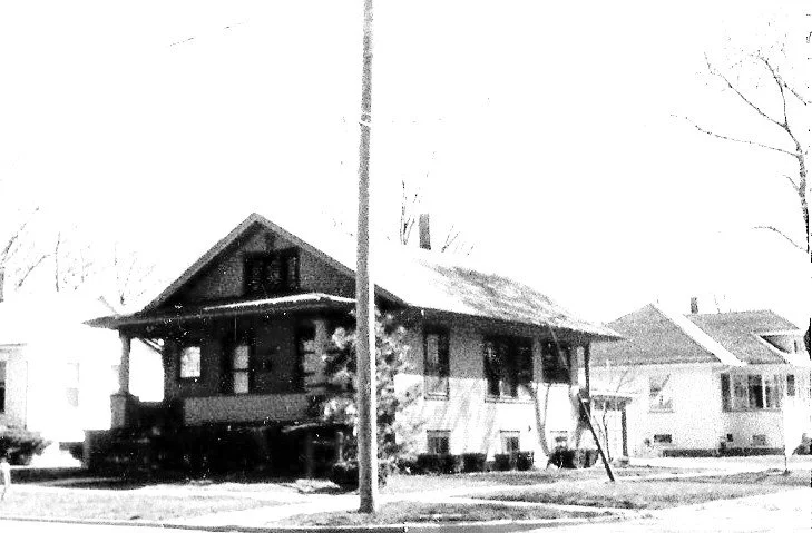 Black-and-white photo of a single-story bungalow-style house on a residential street with a utility pole in the foreground.