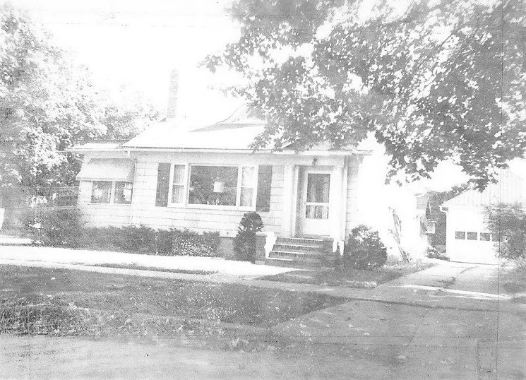 Black-and-white photo of a one-story house with front steps and a large picture window, a tree partially shading the yard, and a detached garage to the right.