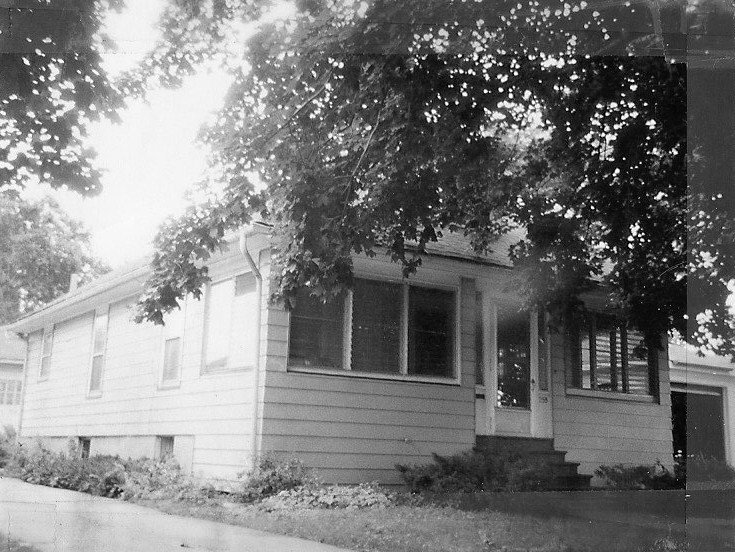 Black-and-white photo of a single-story suburban house with a screened front porch, concrete steps, and large overhanging tree branches.
