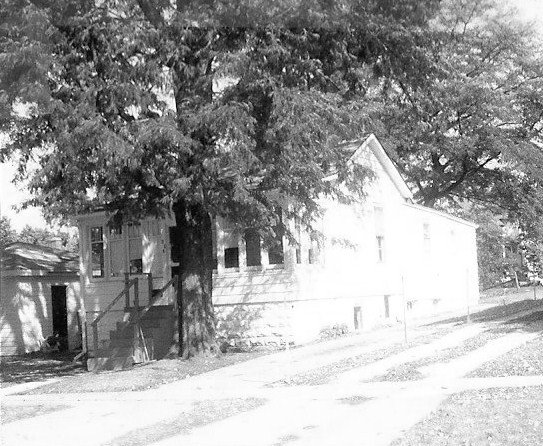 Black-and-white photo of a single-story house with a small front porch, partly hidden by a large tree.