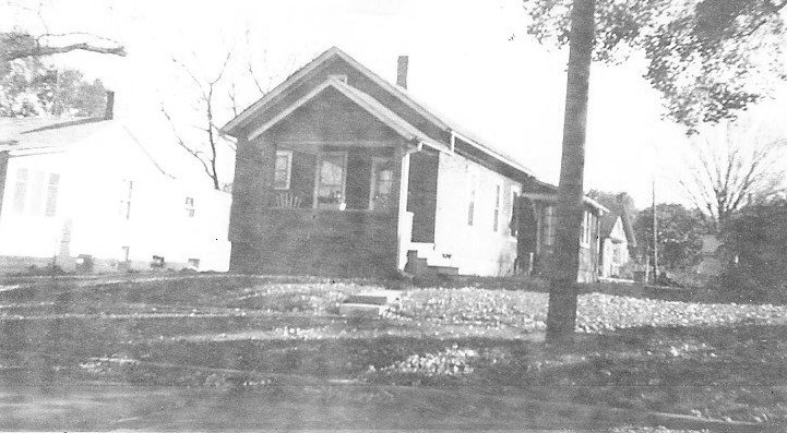 Black-and-white photo of a small single-story bungalow with an enclosed front porch, a tree in the front yard, and neighboring houses.