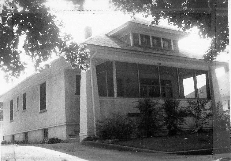Black-and-white photo of a two-story house with an enclosed front porch, shrubs along the foundation, and tree branches framing the roof.