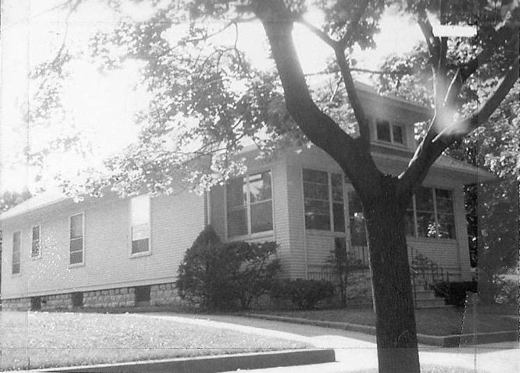 Black-and-white photo of a single-story house with an enclosed front porch, stone foundation, shrubs, and a large tree in the foreground.
