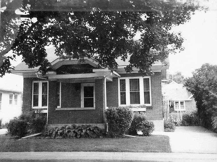 Black-and-white photo of a one-story brick bungalow with a small covered front porch, large overhanging tree, shrubs along the foundation, and a driveway to the right.