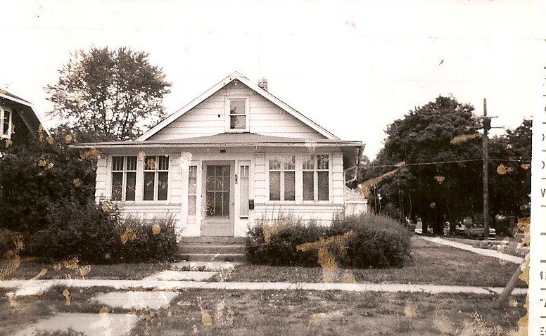 Black-and-white photo of a small single-story bungalow with an enclosed front porch, overgrown shrubs, and a sidewalk and street in front.