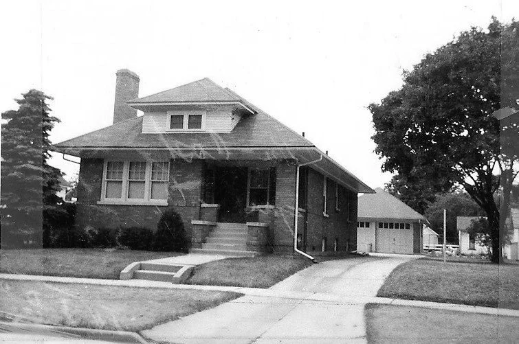 Black-and-white photo of a single-story brick bungalow with front steps, a driveway leading to a detached garage, and a large tree on the right.