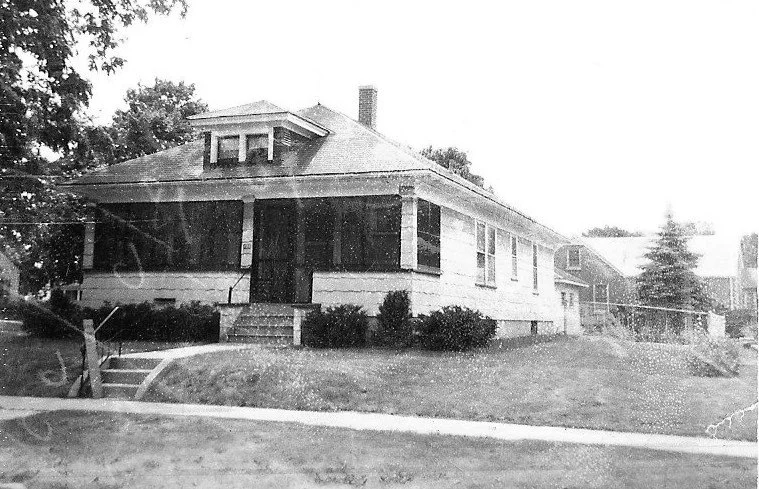Black-and-white photo of a single-story bungalow with a wide screened front porch, front steps, low shrubs, and a sidewalk in the foreground.