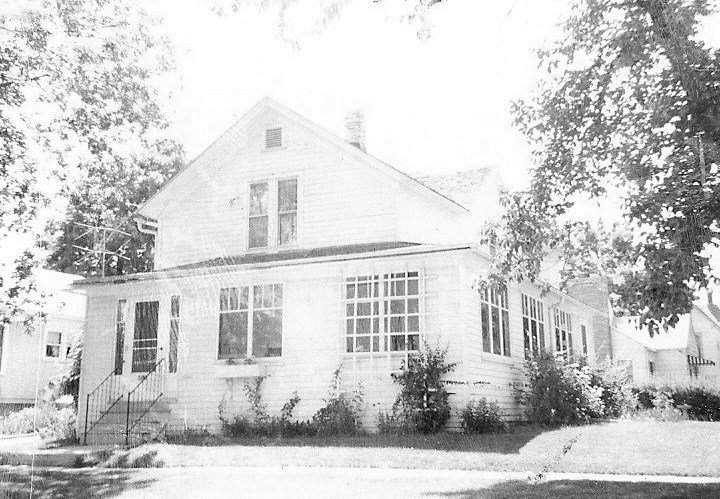 Black-and-white photo of a two-story wood-frame house with an enclosed front porch, entry steps, and trees around it.
