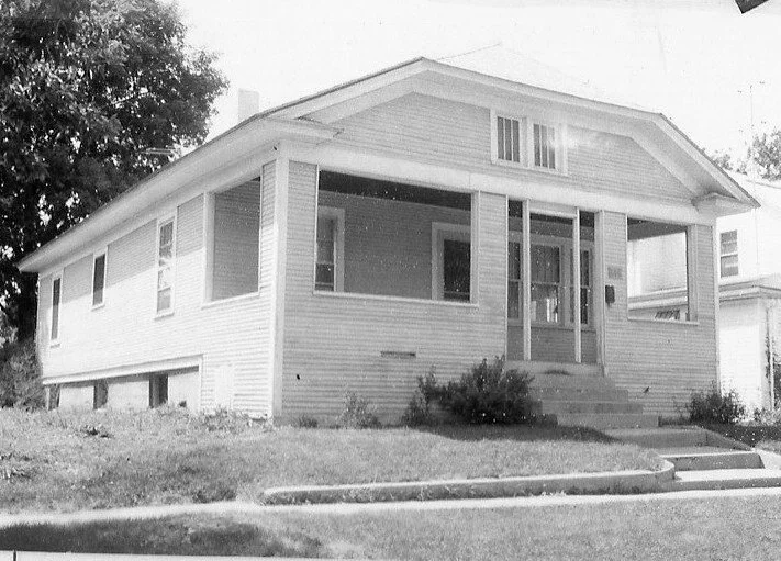 Black-and-white photo of a single-story bungalow with a covered front porch, steps up to the entrance, and a small grassy lawn in front.