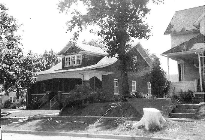 Black-and-white photo of two early 20th-century houses on a tree-lined street, one with a covered front porch and a large tree in front and a fresh tree stump at the sidewalk.