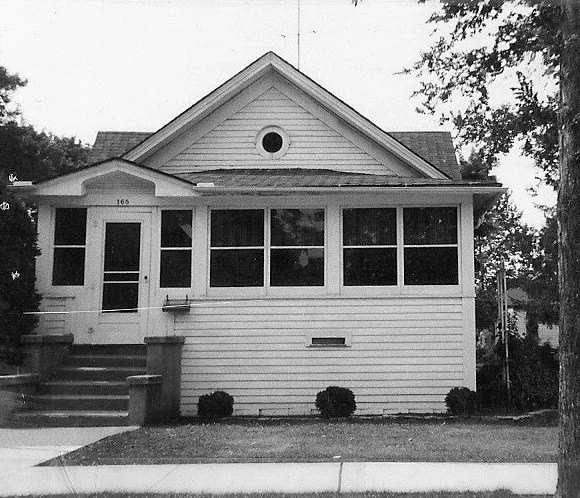 Black-and-white photo of a small single-story bungalow with an enclosed front porch, six-pane windows, concrete steps to a door marked “165,” and low shrubs along the foundation.