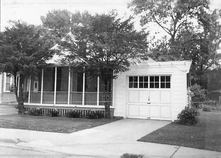 Black-and-white photo of a single-story house with a screened front porch, two small trees, and a driveway leading to a single-car garage.