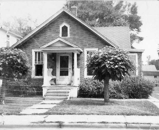 Black-and-white photo of a small gabled bungalow with a front porch, two rounded shade trees in the yard, and a walkway leading to the steps.