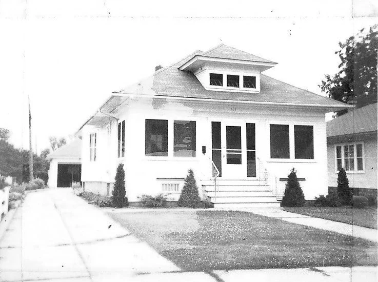 Black-and-white photo of a single-story bungalow with a screened front porch, steps leading to the entrance, a driveway to a detached garage, and small shrubs in the yard.
