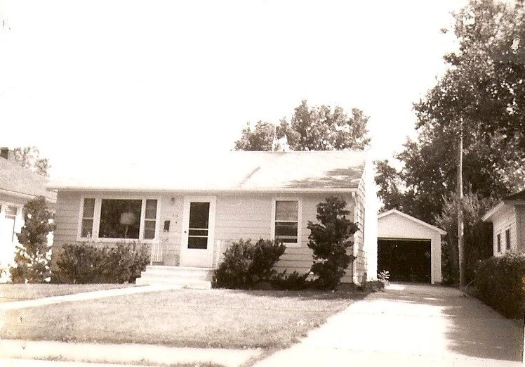 Black-and-white photo of a single-story suburban house with a front lawn, shrubs, a driveway leading to a detached garage at right, and trees in the background.