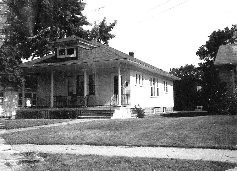 Black-and-white photo of a single-story bungalow with a covered front porch, steps, and a grassy lawn.