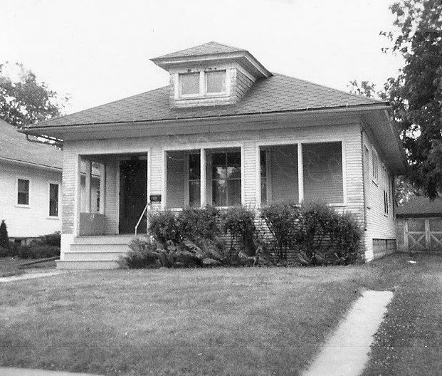 Black-and-white photo of a single-story bungalow with a front porch, dormer window, shrubs across the front, and a lawn with a sidewalk.
