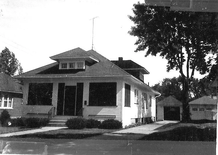 Black-and-white photo of a single-story bungalow with a covered front porch, steps, driveway leading to a detached garage, and a large tree at the right.