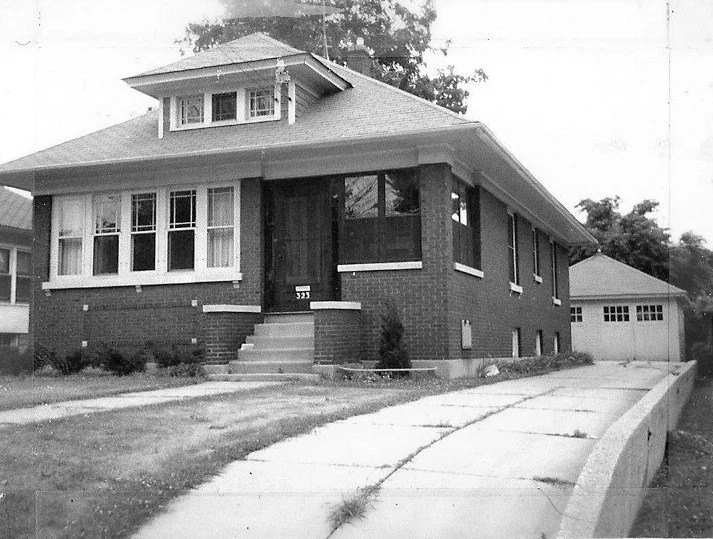 Black-and-white photo of a single-story brick bungalow with front steps, a sloped driveway leading to a detached garage, and the house number "323" visible by the entrance.