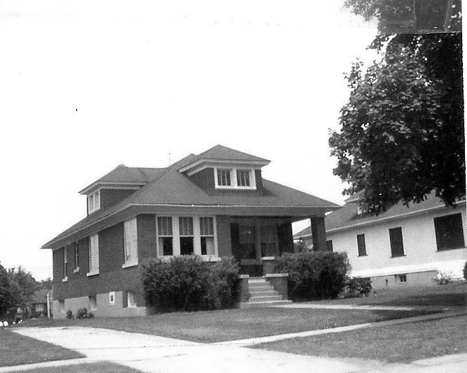Black-and-white photo of a single-story brick bungalow with a covered front porch, dormer windows, shrubs flanking the steps, and a sidewalk in front.