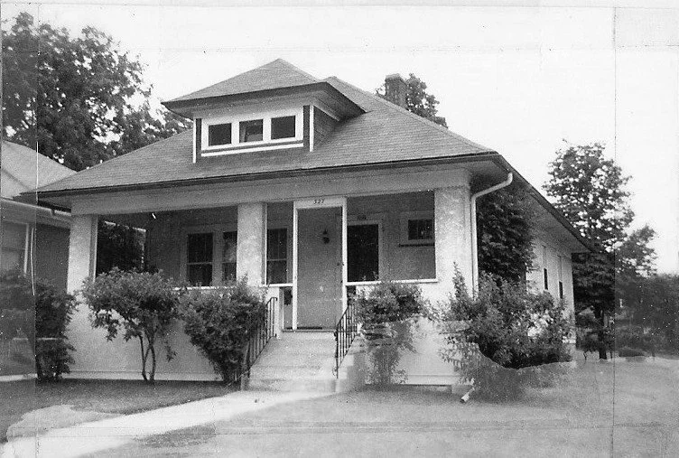 Black-and-white photo of a single-story bungalow with a covered front porch, central entrance reached by steps, low-pitched roof with a dormer, and shrubs lining the front yard.