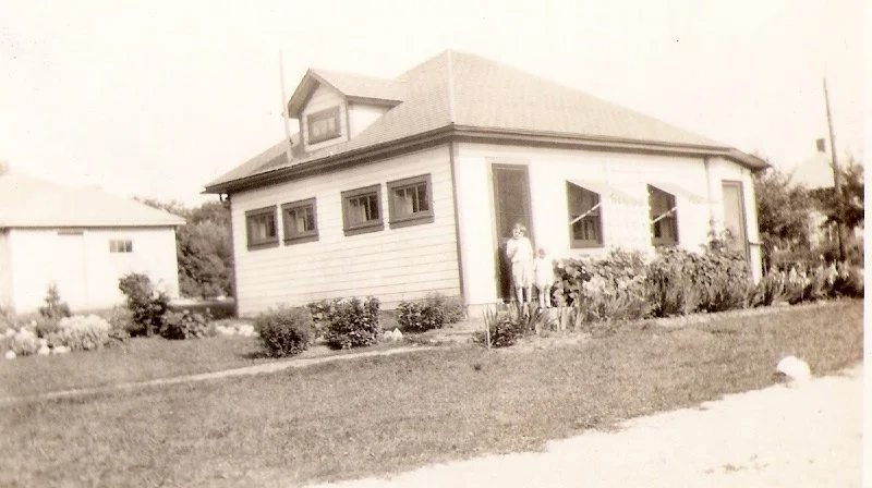 Vintage black-and-white photo of a small one-story house with a front porch, garden beds, and a person standing by the entrance.