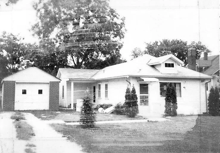 Black-and-white photo of a single-story bungalow with a detached garage, small front yard, driveway, and several shrubs.
