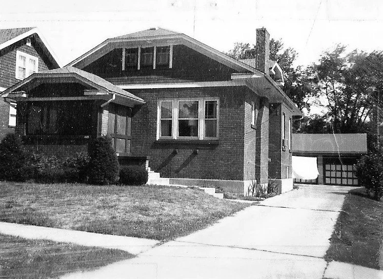 Black-and-white photo of a one-story brick bungalow with a covered front porch, lawn, driveway, and detached garage.