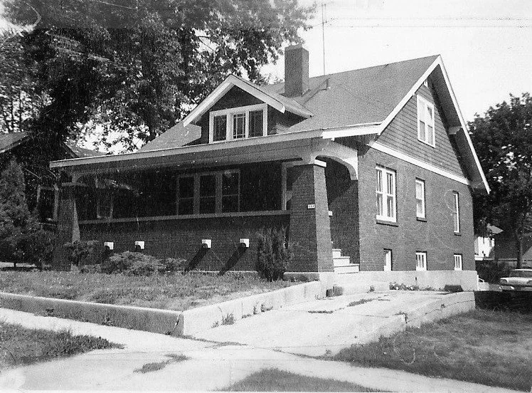 Black-and-white photo of a one-and-a-half-story brick bungalow with a covered front porch, dormer window, and concrete driveway.