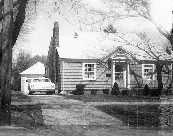 Black-and-white photo of a single-story suburban house with a front porch, driveway leading to a detached garage, and a car parked beside it, leafless trees in the yard.