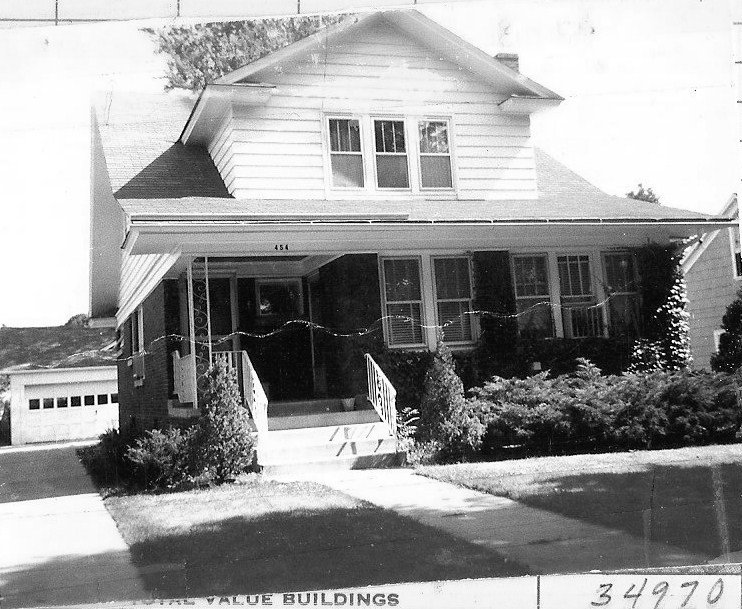 Black-and-white photo of a two-story suburban house with a covered front porch, steps and railings, shrubs in the yard, and a detached garage; "454" visible above the porch.