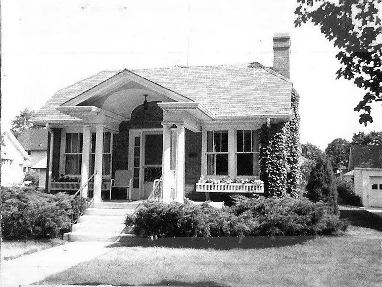Black-and-white photo of a one-story brick bungalow with a covered front porch, columns, chimney, window boxes, shrubs, and a sidewalk leading to the entrance.