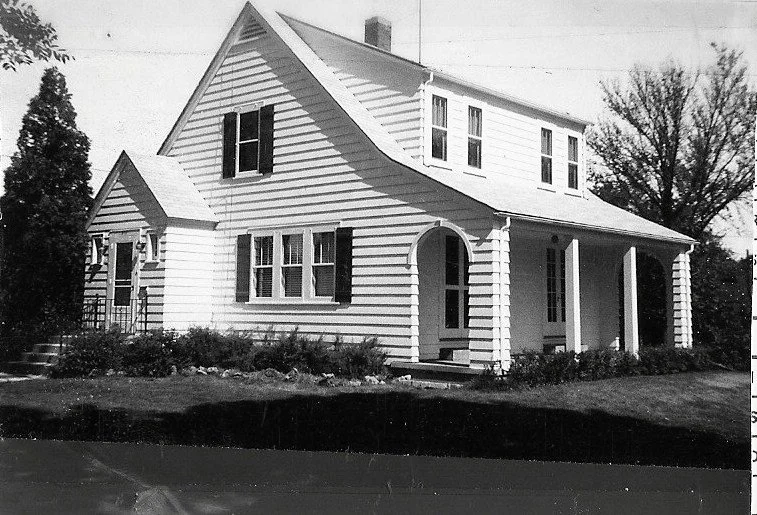Black-and-white photo of a two-story white clapboard house with an arched front entry, covered porch, lawn, and surrounding trees.