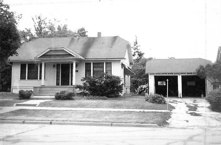 Black-and-white photo of a single-story bungalow with a front porch, shrubs and lawn, and a detached two-car garage to the right.