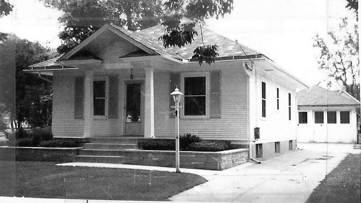 Black-and-white photo of a single-story bungalow with a front porch and steps, a lamp post on the lawn, and a driveway leading to a detached garage.