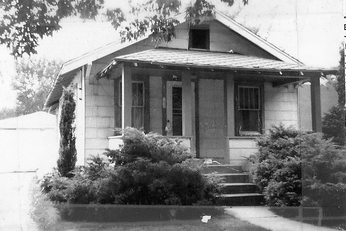 Black-and-white photo of a small single-story bungalow with a covered front porch, concrete steps and dense shrubs framing the entrance.