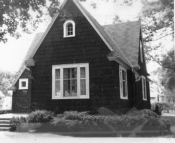 Black-and-white photo of a small steep-roofed cottage-style house with a central bay window, arched dormer window, and low shrubs along the front.