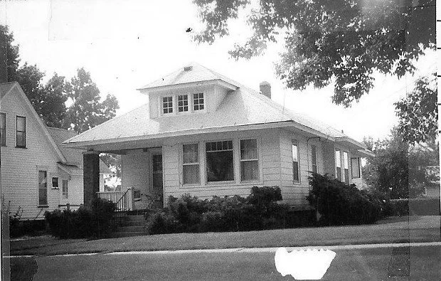 Black-and-white photo of a single-story bungalow with a wide front porch, dormer window, and shrubs along the front yard.