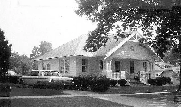 Black-and-white photo of a single-story bungalow with a front porch and a vintage car parked beside it, tree branches overhead.