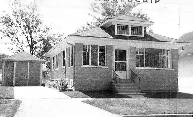 Black-and-white photo of a one-story brick bungalow with an enclosed front porch, center steps, and a detached garage.
