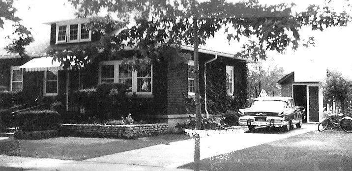Black-and-white photo of a brick bungalow with trees and shrubs, a vintage car parked in the driveway next to a small garage.