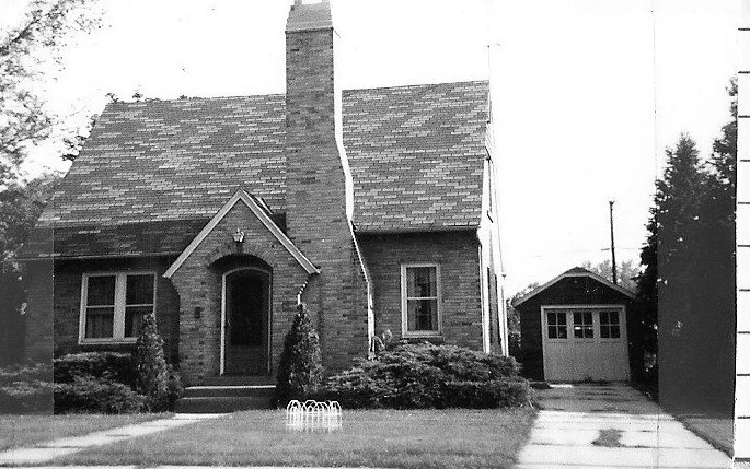 Black-and-white photo of a small brick cottage with a steep shingled roof and central chimney, arched front entry, shrubs in the yard, and a detached garage to the right.