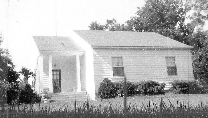 Black-and-white photo of a small single-story white house with a covered front porch, steps, two front windows, shrubs along the foundation and a grassy lawn in the foreground.